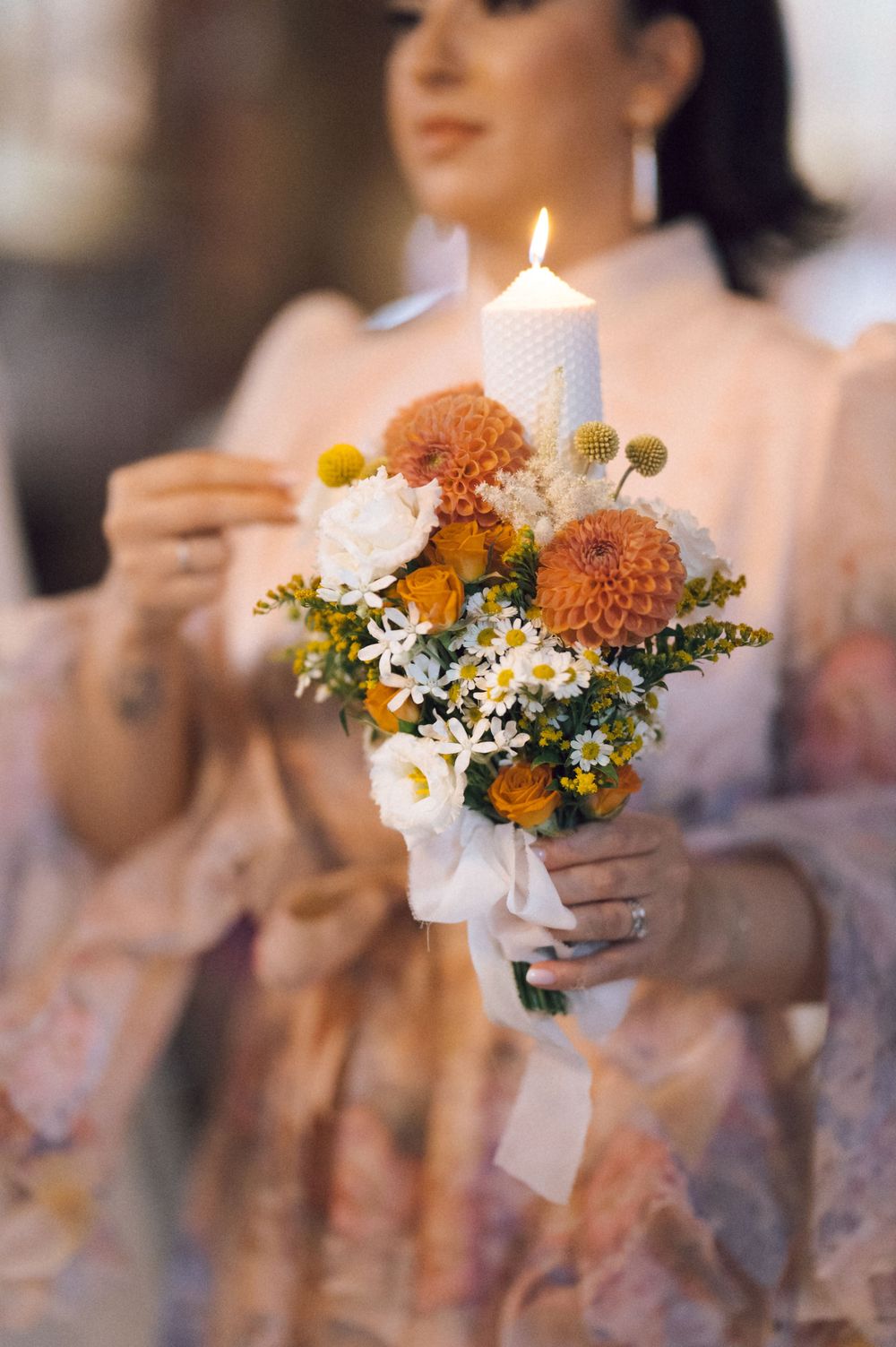 Woman in pink dress holds a floral arrangement with a lit white candle at a wedding ceremony.