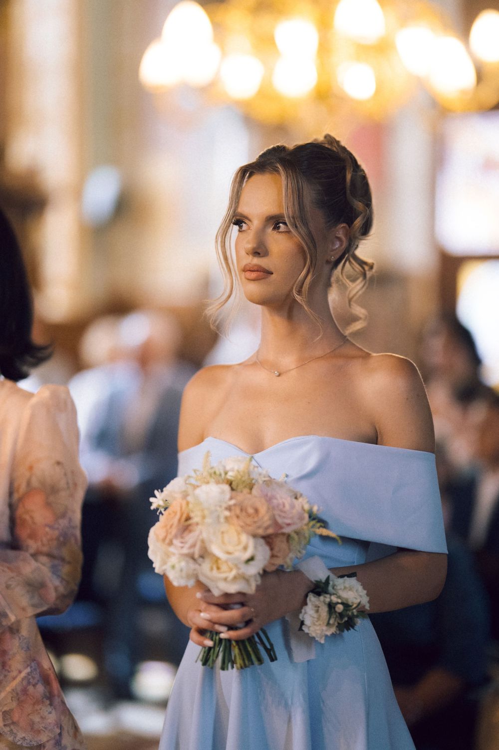 Elegant bride in off-shoulder blue gown holds a pastel rose bouquet inside a grand church.
