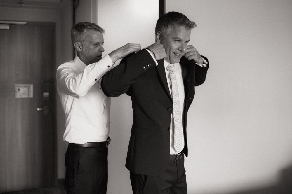 Two men in suits preparing for a wedding, one helping the other adjust his jacket collar in black and white.