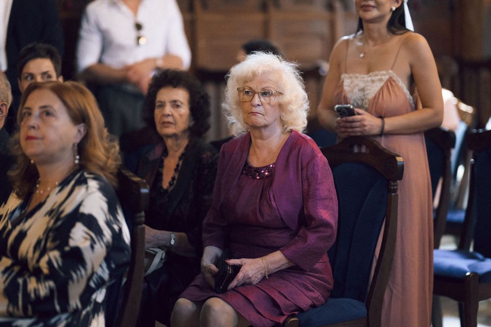 Elegantly dressed women seated at a formal event, featuring a blonde woman in burgundy dress with glasses.