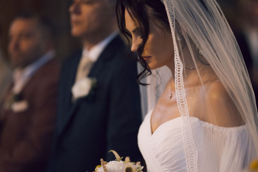 Bride in white gown and veil holding bouquet, bowing head during wedding ceremony in church.