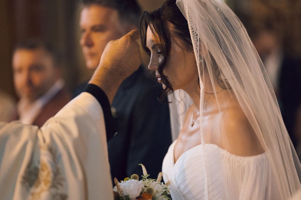Bride in white veil receives anointing from priest during Catholic wedding ceremony while holding bouquet.