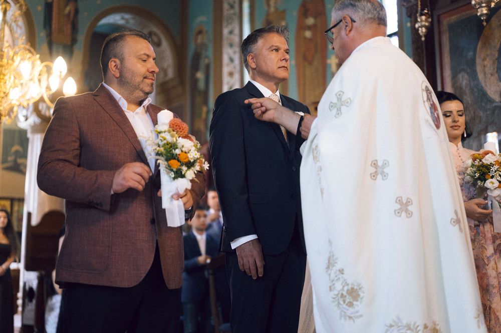 Men in formal attire participating in an Orthodox Christian church ceremony with a priest in white robes.