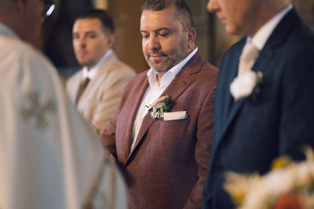Groom in brown plaid suit with boutonniere stands solemnly during wedding ceremony with groomsmen in background.