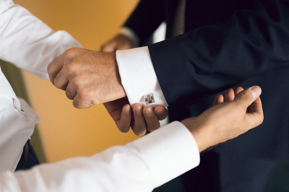 Man in black suit getting cufflinks fastened on white dress shirt sleeves by another person.