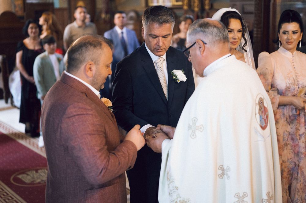Priest officiating a wedding ring exchange ceremony with groom in brown suit at a church altar.