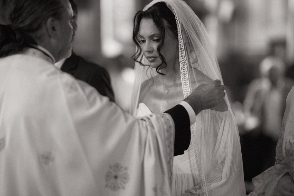 Black and white wedding ceremony photo of bride in veil receiving blessing from priest at church altar.
