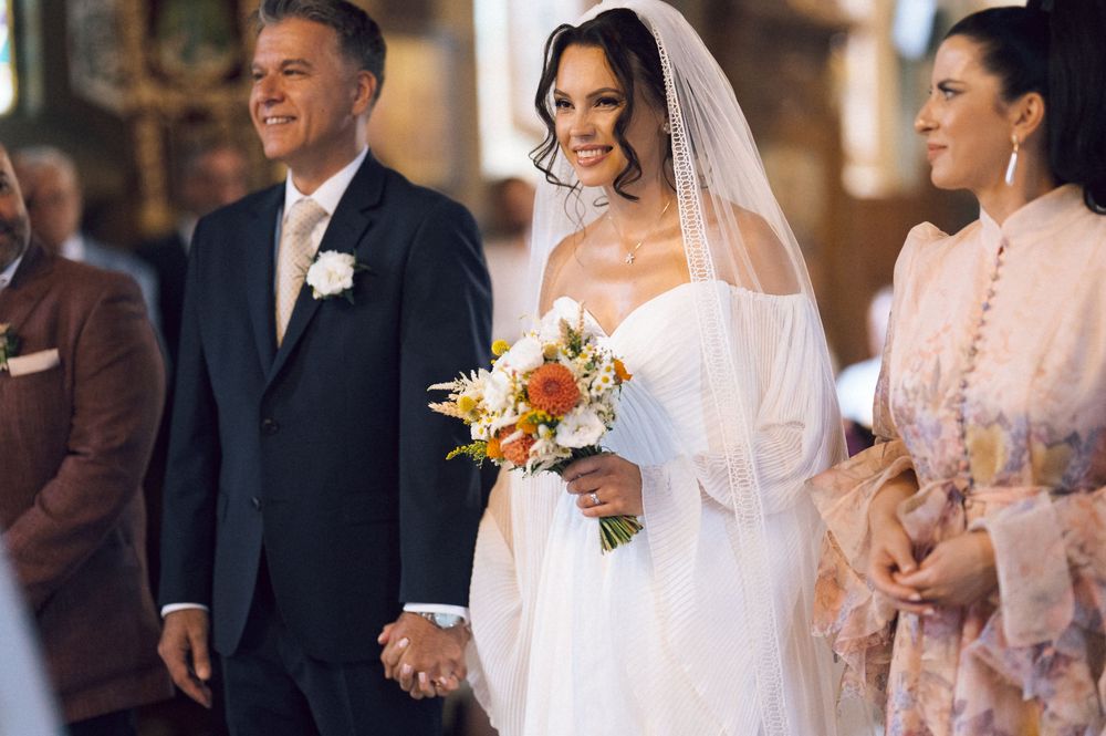 Bride in white gown and veil holds bouquet while walking down church aisle with older man in dark suit.