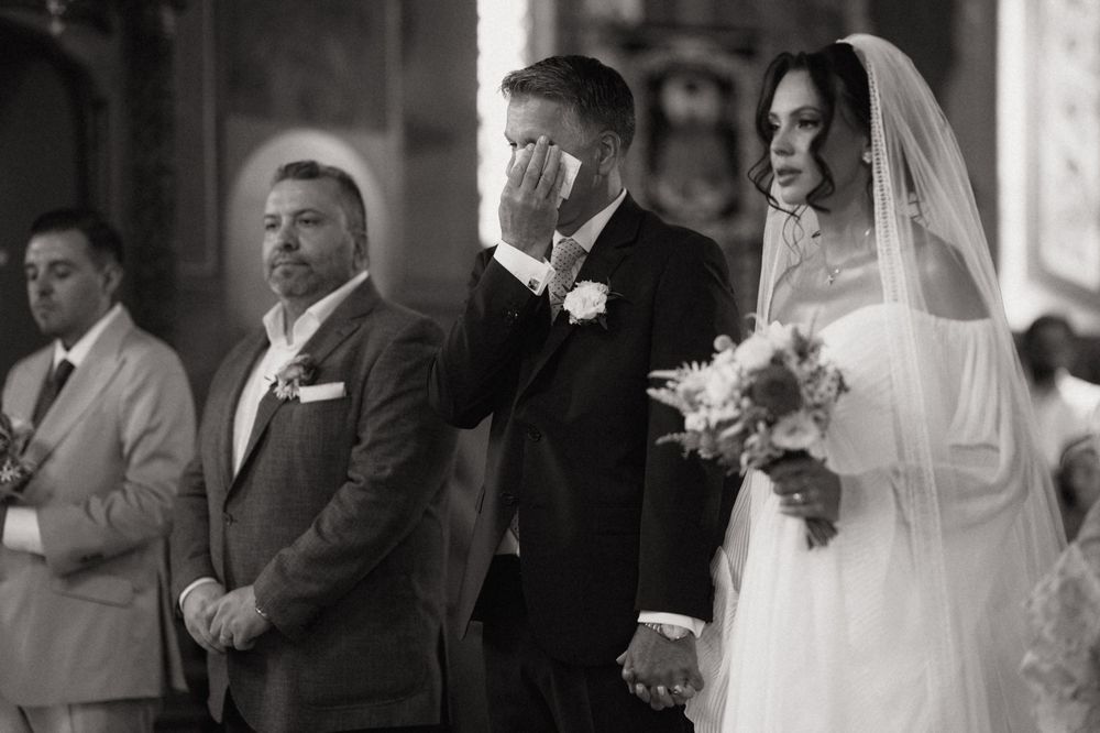 Emotional groom wiping tears during wedding ceremony with bride holding bouquet in black and white photo.