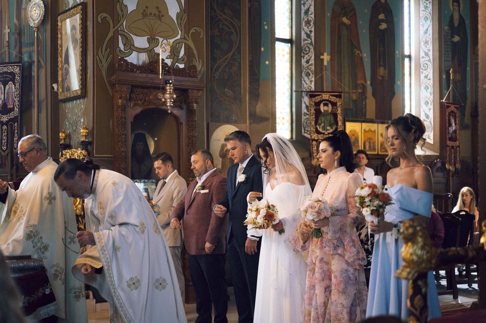 Orthodox church wedding ceremony with bride in white gown, groom, bridesmaids, and priest at ornate altar.