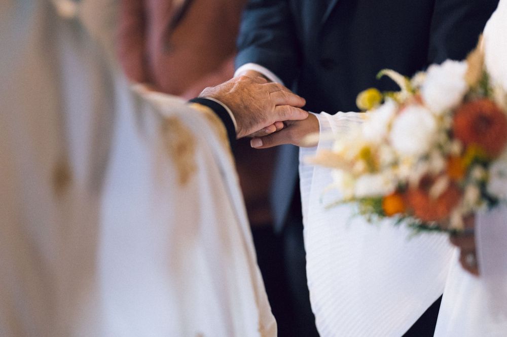 Close-up of groom holding bride's hand during wedding ceremony, with bridal bouquet of white and orange flowers.