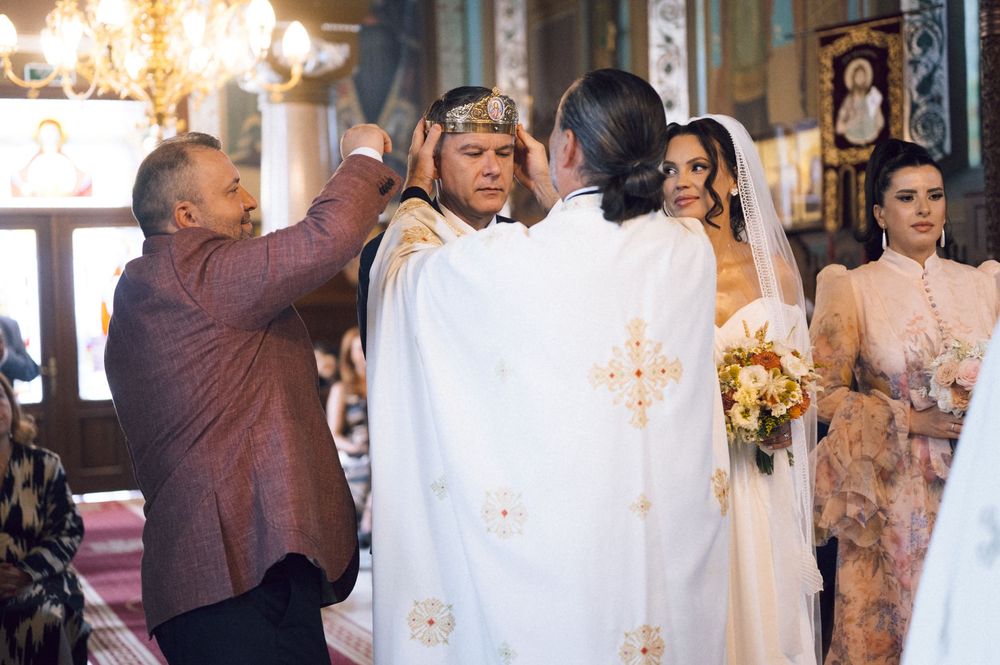 Orthodox wedding ceremony with priest placing crown on groom, bride holding bouquet, inside ornate church.