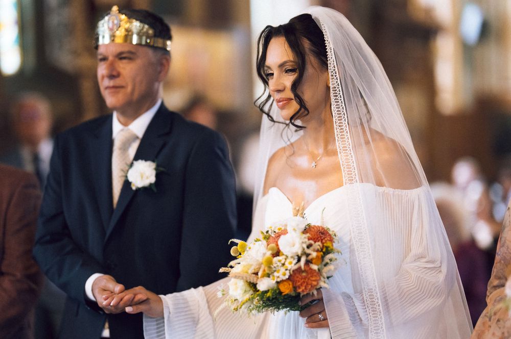 Bride in white gown and veil holds colorful bouquet beside suited man wearing gold crown in church.