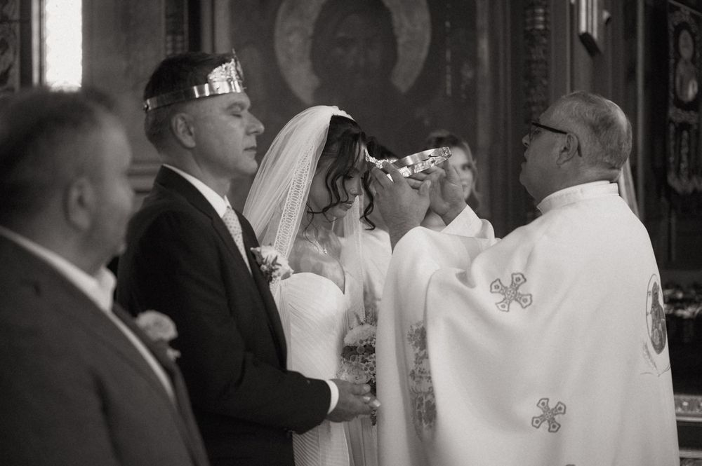 Black and white photo of a couple during an Orthodox wedding ceremony with a priest placing crowns on their heads.