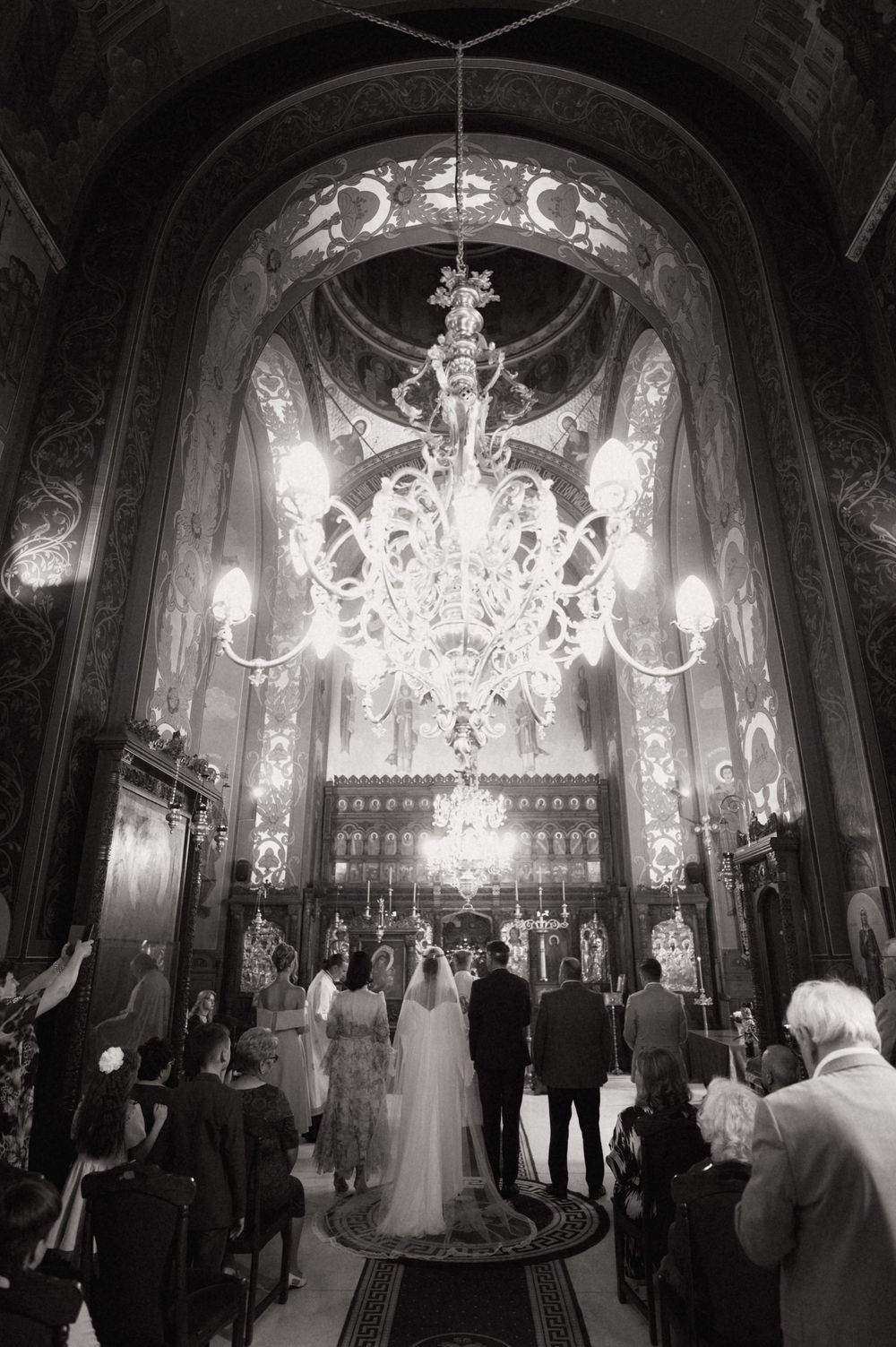 Black and white wedding ceremony photo inside ornate church with chandelier, bride and groom at altar.