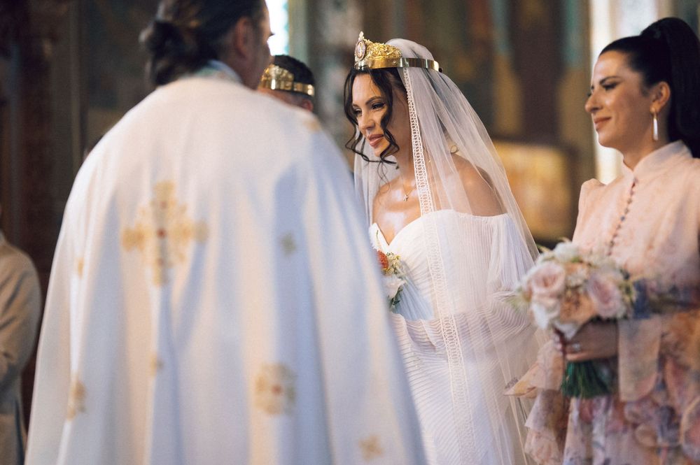 Bride in white gown and crown veil smiles at Orthodox priest during traditional church wedding ceremony.