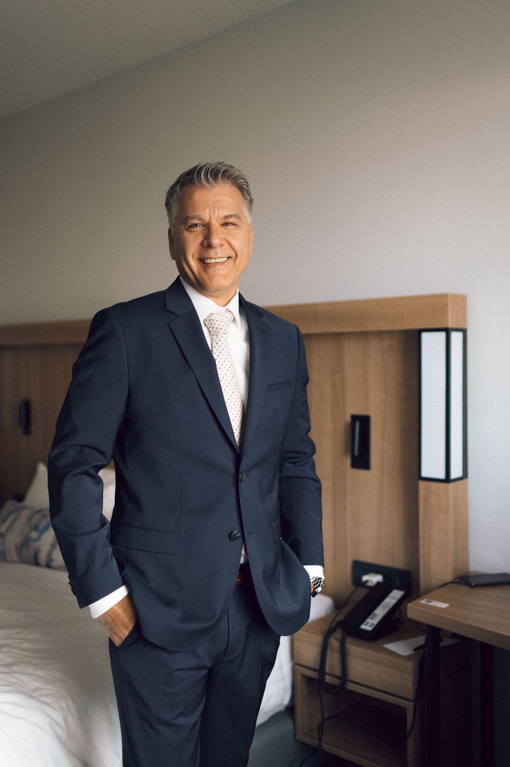 A smiling middle-aged businessman in a navy suit stands confidently in a modern hotel room with hands in pockets.