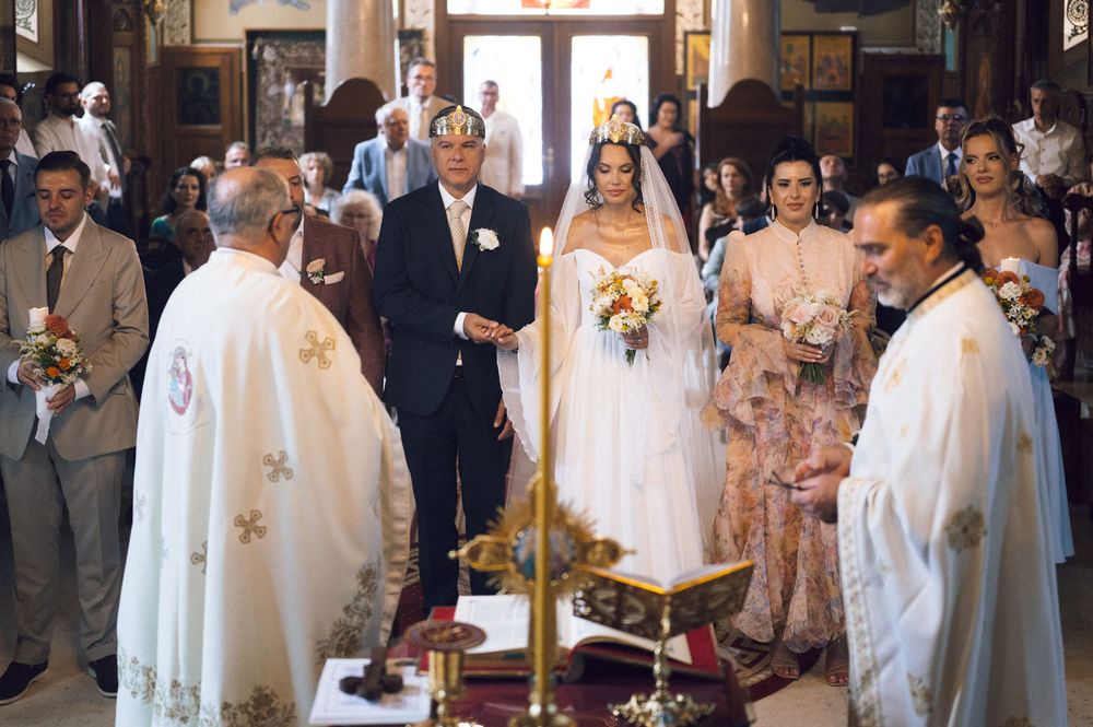 Bride walks down the aisle with escort during a traditional Orthodox church wedding ceremony with priests officiating.