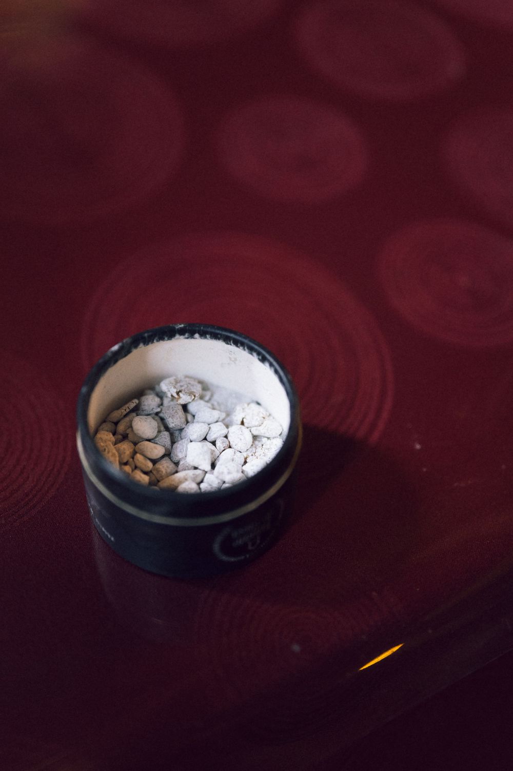 Close-up of an open tin container filled with small white powder-coated nuts on a dark red patterned surface.