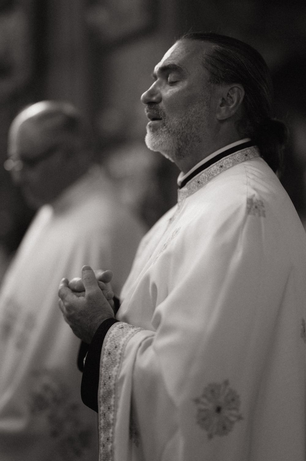 Black and white photo of a priest in white vestments praying with eyes closed during a religious ceremony.