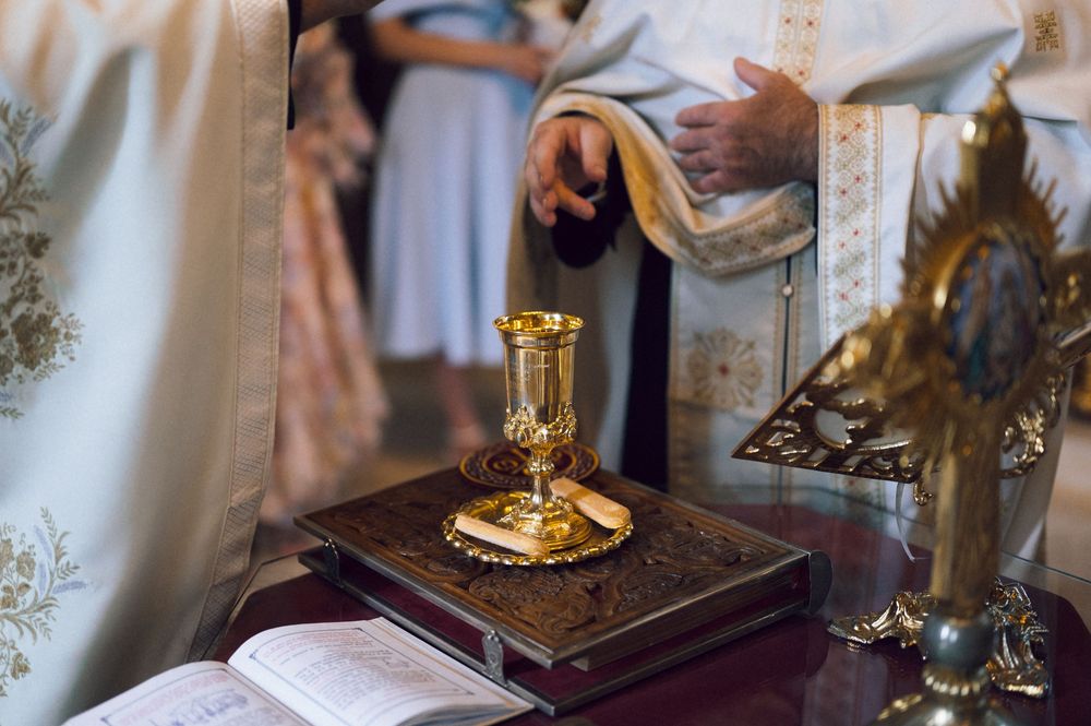 Golden chalice and ornate Bible on altar during Orthodox Christian liturgy ceremony with priest in white vestments.