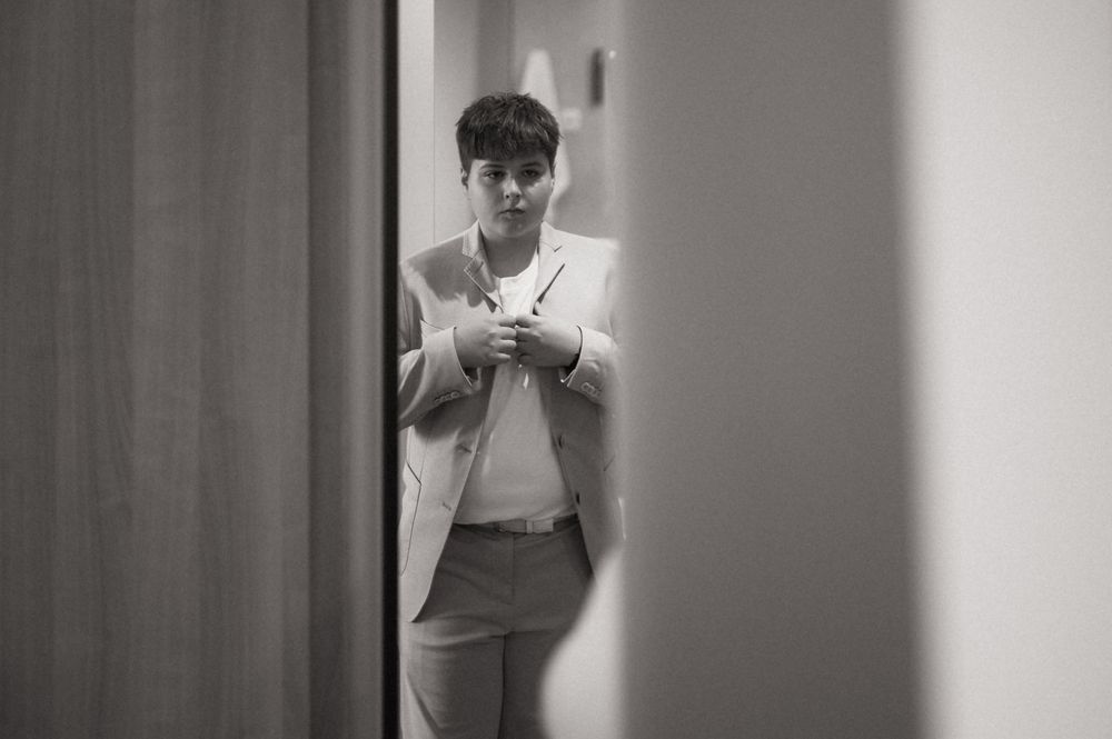 Black and white photo of a person in a light blazer adjusting their collar in a hallway.