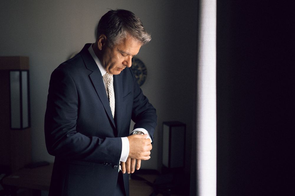 Businessman in dark suit checking wristwatch in dimly lit room, looking down with focused expression.