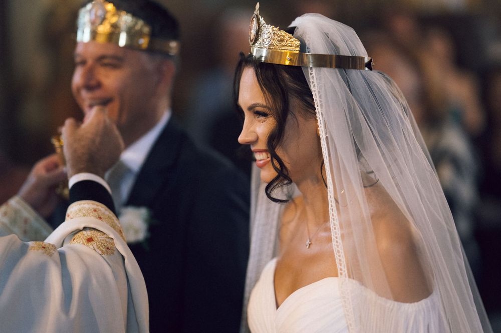 Smiling bride wearing a gold crown and white veil during a wedding ceremony beside a priest in golden vestments.
