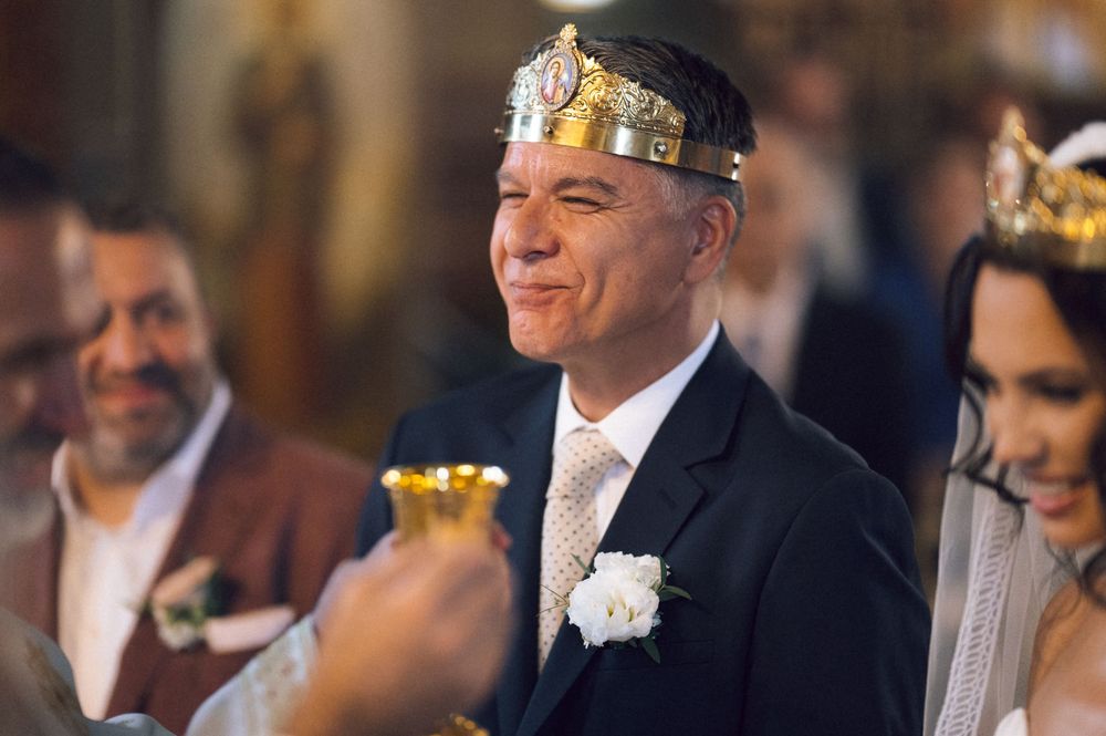 Groom wearing gold crown smiles during Orthodox wedding ceremony in ornate church.