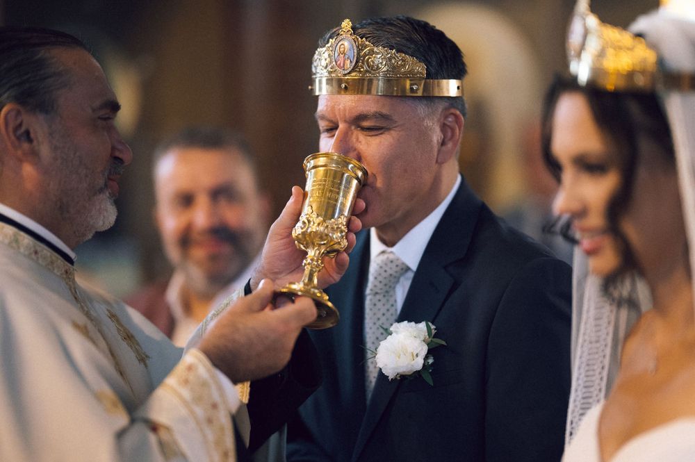 Groom drinks from golden chalice during Orthodox wedding ceremony, wearing crown, with bride and priest beside him.