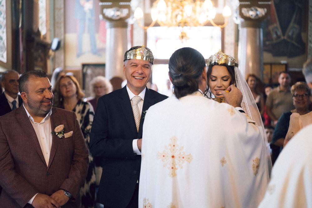 Orthodox wedding ceremony with crowned bride and groom smiling before a priest in an ornate church.
