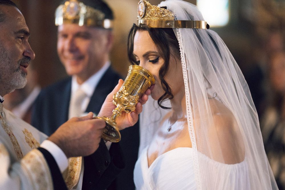 Bride drinking from ornate golden chalice during Orthodox wedding ceremony with priest and guests.