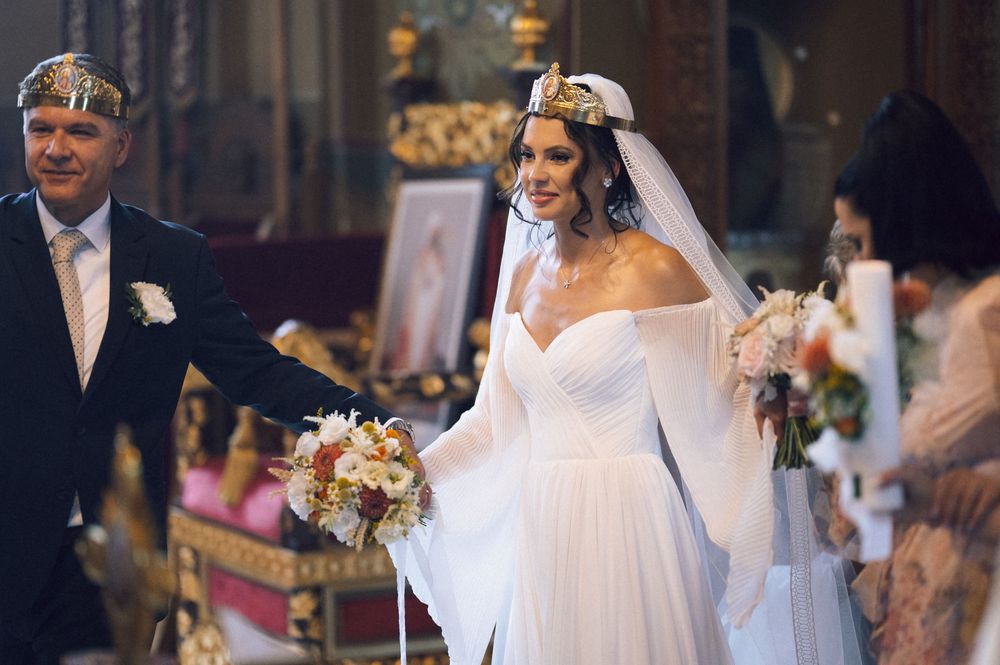 Elegant bride in white off-shoulder gown holding bouquet during Orthodox church wedding ceremony.