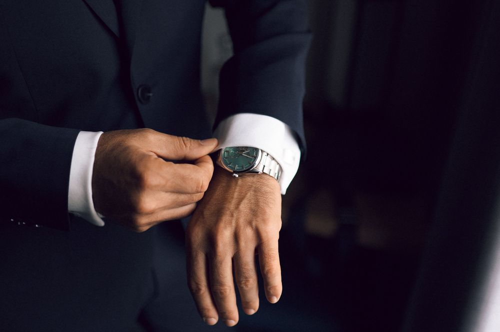 Man in dark suit adjusting silver wristwatch, showcasing elegant formal attire and luxury timepiece.