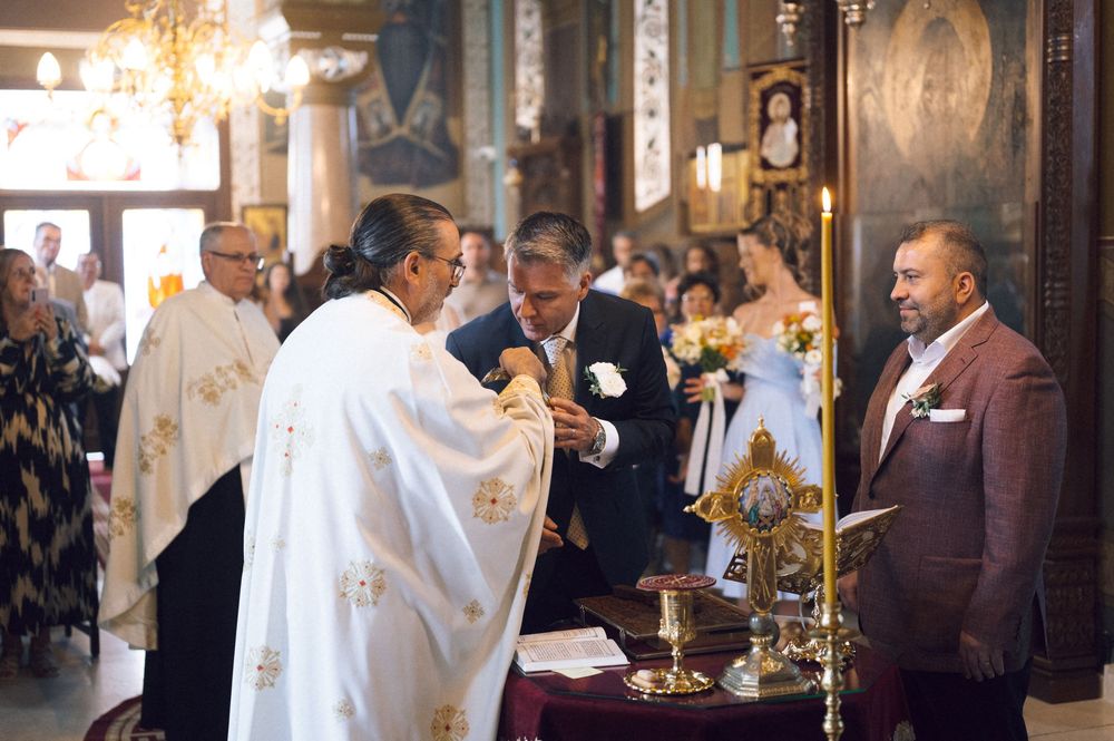 Groom kissing priest's hand during Orthodox wedding ceremony in ornate church with guests watching.