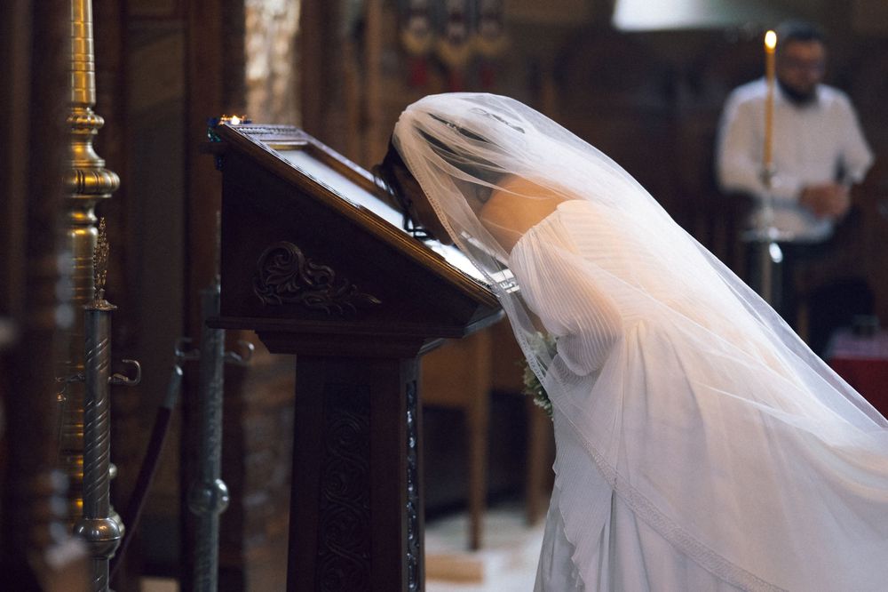 Bride in white veil bowing head in prayer at church altar during wedding ceremony.