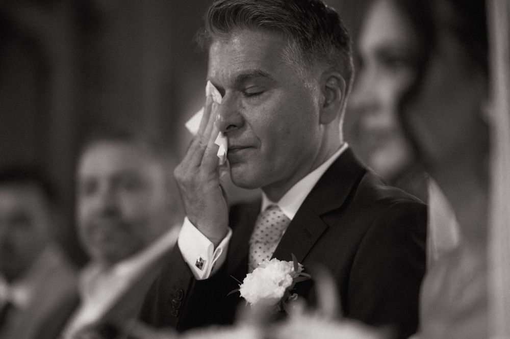 Emotional groom wiping tears at wedding ceremony in black and white photo.