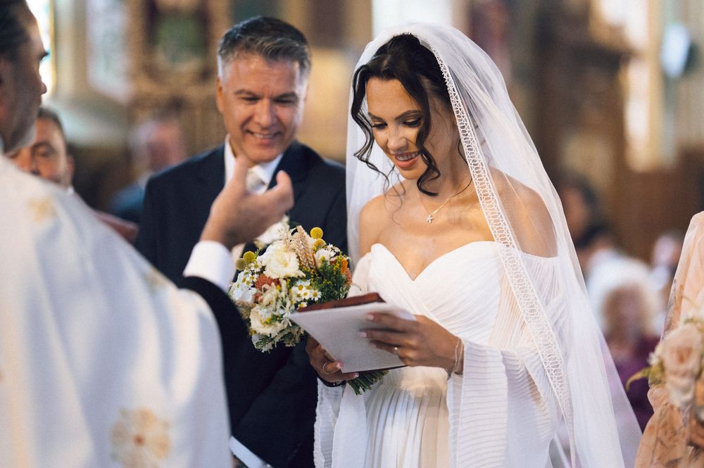 Bride in white gown and veil reads vows at church altar, holding bouquet, with smiling groom beside her.