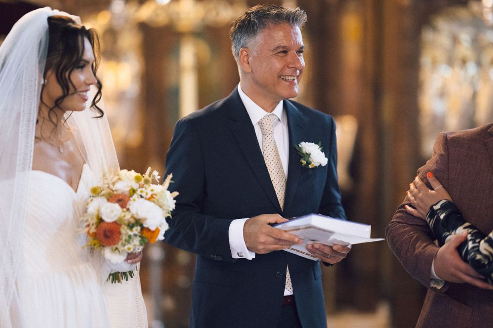 Smiling groom in navy suit holds booklet beside bride in white veil holding orange and white bouquet at wedding ceremony.