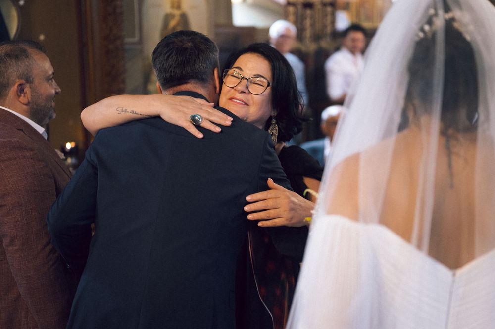 Emotional wedding guest hugging groom during ceremony, bride in white veil visible in background.