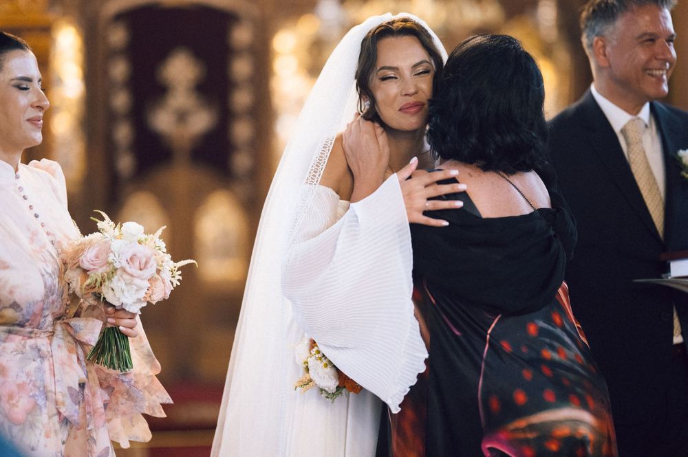 Bride in white gown hugging guest at church wedding ceremony while holding bouquet of blush pink flowers.