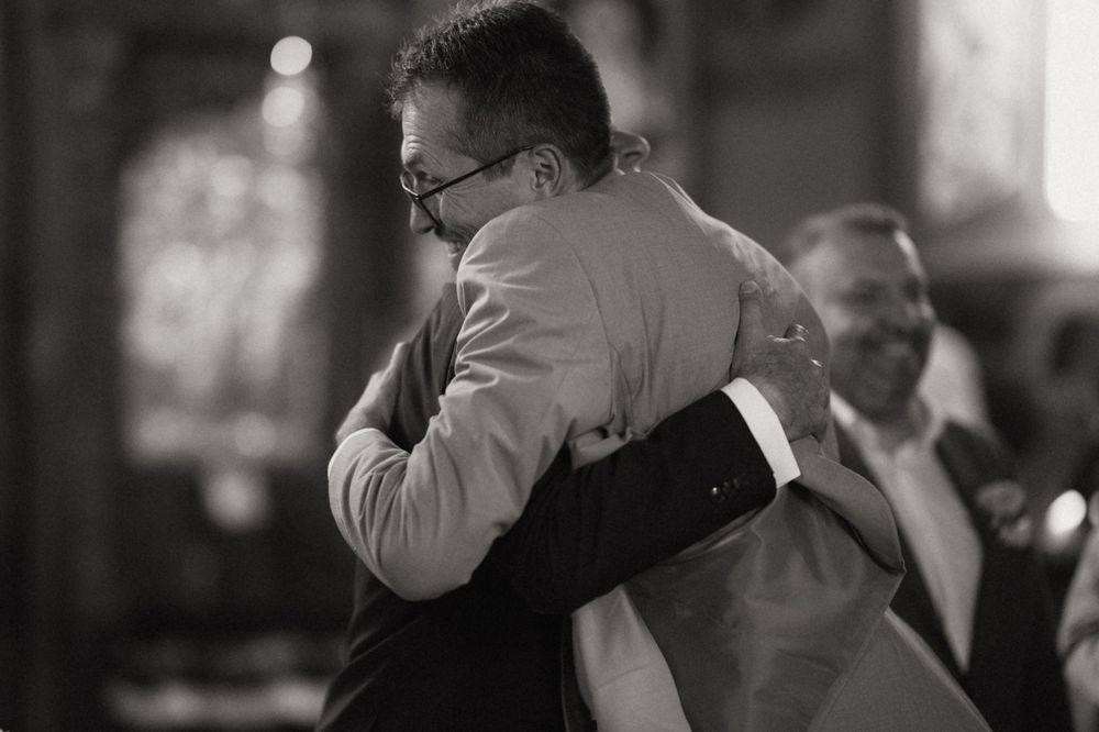 Two men share an emotional hug at a wedding ceremony in a church, captured in black and white photography.