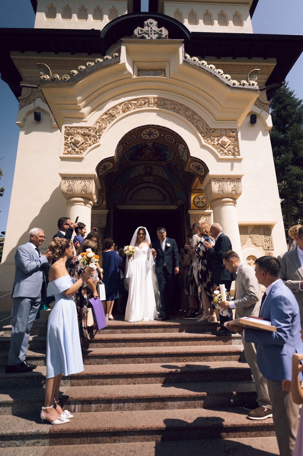 Bride and groom exit ornate church entrance on steps, surrounded by wedding guests celebrating outdoors.
