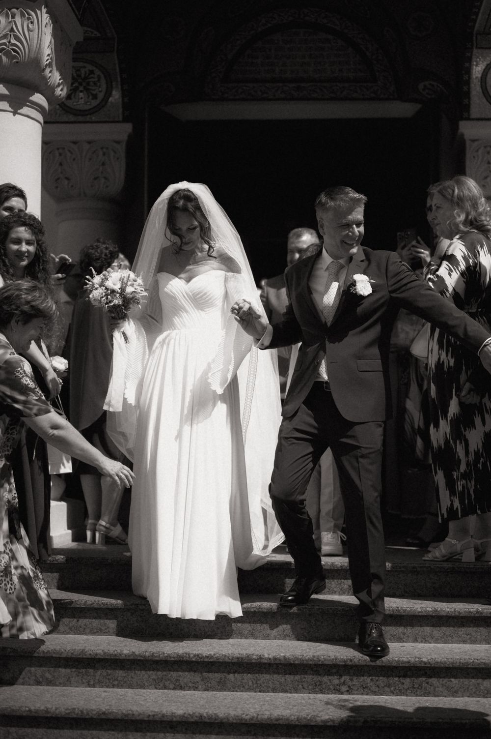 Black and white photo of bride and groom descending church steps, surrounded by smiling wedding guests.