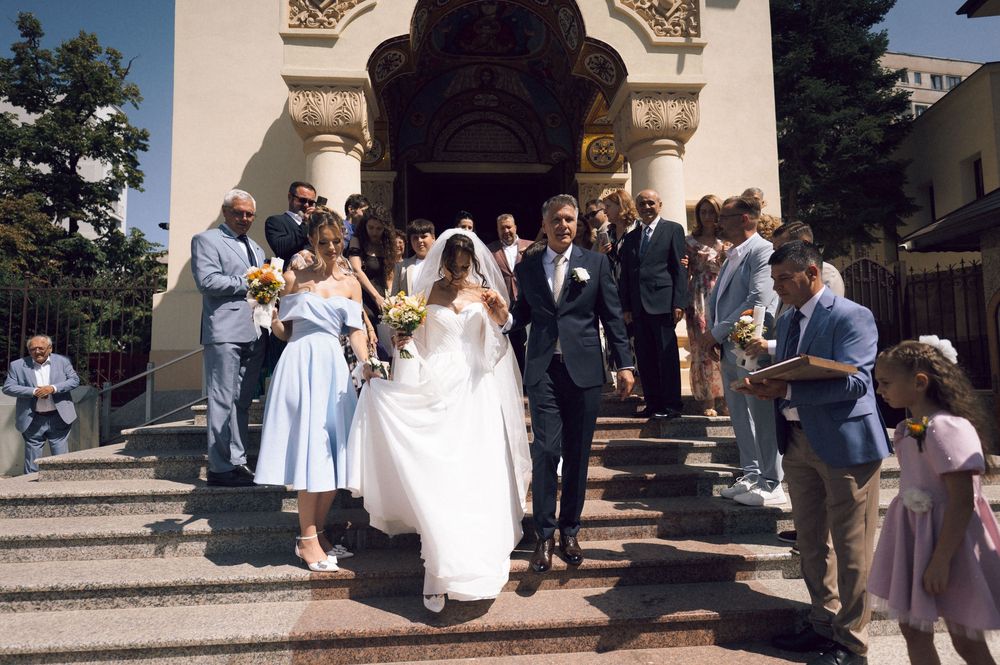 Bride and groom exit a church surrounded by wedding guests on sunny steps after their ceremony.
