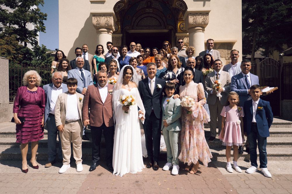 Large wedding group photo with bride and groom surrounded by family and guests on church steps.