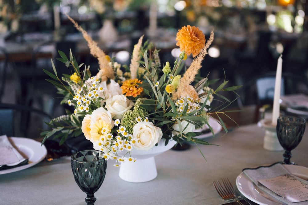 Elegant wedding centerpiece with orange dahlias, white roses, and greenery in a white vase on a reception table.