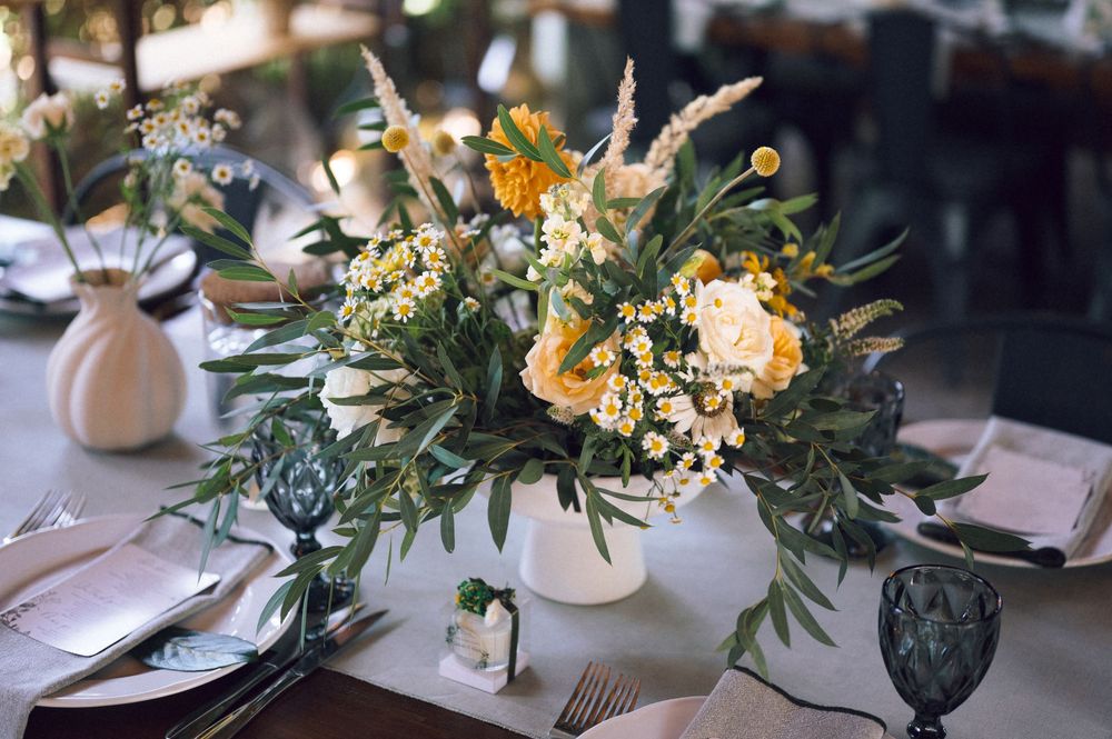 Rustic wedding centerpiece with yellow flowers, white roses, daisies, and lush greenery in a white vase on an elegant table.