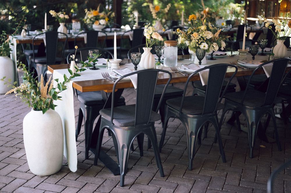 Elegant outdoor wedding reception with dark metal chairs, wooden tables, floral centerpieces, and large white vases on brick patio.