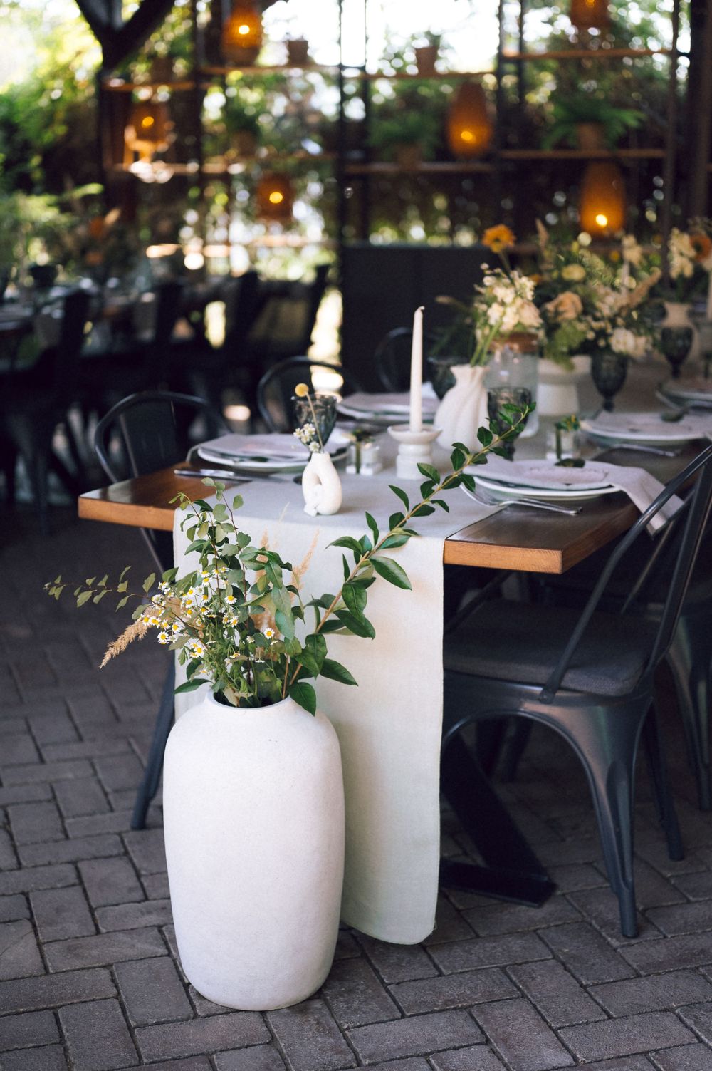 Elegant outdoor wedding reception table with white ceramic vases, greenery, candles, and metal chairs on brick patio.
